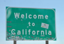 Green 'Welcome to California' road sign covered with various stickers against a clear blue sky.